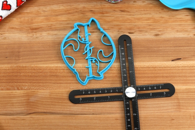 May include: A blue plastic cookie cutter shaped like two cats facing each other. The cutter is on a wooden surface with a black ruler.