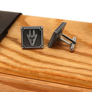 May include: A pair of silver cufflinks with a square design featuring a stylized symbol. The cufflinks are displayed in a wooden box with a leather strap.