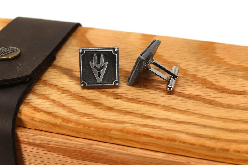 May include: A pair of silver cufflinks with a square design featuring a stylized symbol. The cufflinks are displayed in a wooden box with a leather strap.