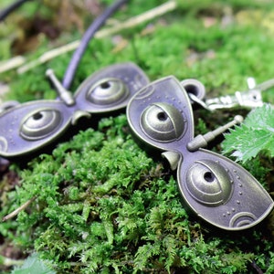 May include: Two bronze-colored butterfly-shaped charms with large, round eyes. One charm is attached to a black cord, and the other is attached to a silver key ring.