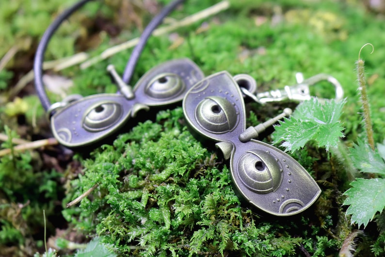 May include: Two bronze-colored butterfly-shaped charms with large, round eyes. One charm is attached to a black cord, and the other is attached to a silver key ring.