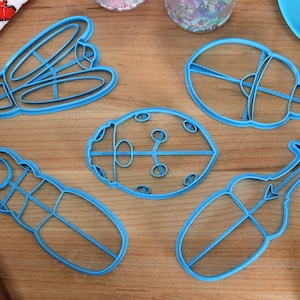 Puede incluir: Cinco cortadores de galletas de plástico azul con forma de insectos. Los cortadores están sobre una superficie de madera. Los cortadores tienen forma de mariquita, escarabajo, libélula y dos tipos diferentes de escarabajos.