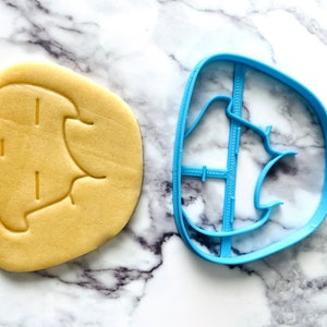 May include: A blue plastic cookie cutter shaped like a cartoon elephant. A yellow cookie is next to the cutter, showing the elephant shape.