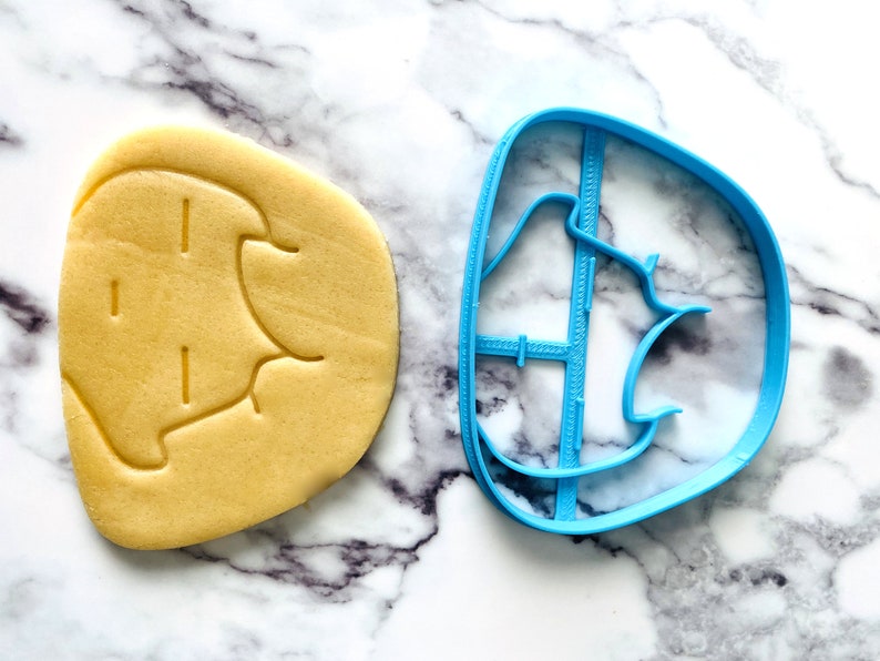 May include: A blue plastic cookie cutter shaped like a cartoon elephant. A yellow cookie is next to the cutter, showing the elephant shape.