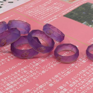May include: A group of 10 purple resin rings with a gold flame design. The rings are faceted and have a translucent appearance.