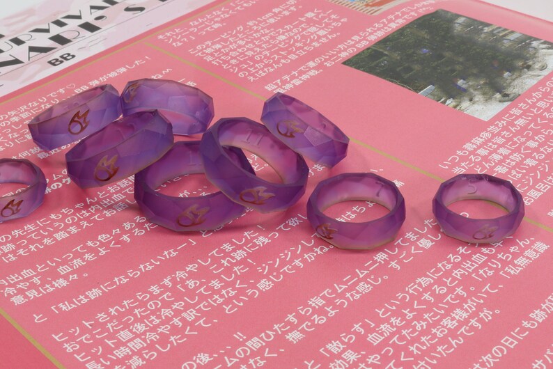 May include: A group of 10 purple resin rings with a gold flame design. The rings are faceted and have a translucent appearance.