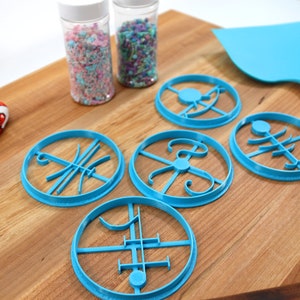 Op de afbeelding: Vier blauwe koekjesvormen met verschillende ontwerpen op een houten snijplank. Twee potten met sprinkles staan op de achtergrond.