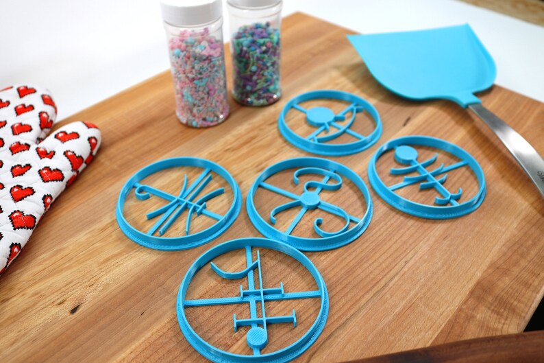 Op de afbeelding: Vier blauwe koekjesvormen met verschillende ontwerpen op een houten snijplank. Twee potten met sprinkles staan op de achtergrond.