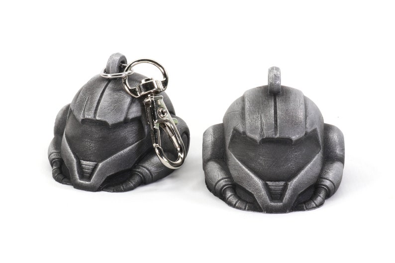 Puede incluir: Un llavero de metal de color plateado que presenta una r&eacute;plica detallada de un casco espacial. El llavero tiene un anillo de metal plateado y un cierre de metal plateado.