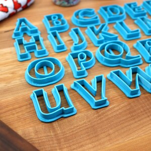 May include: A set of blue plastic alphabet cookie cutters on a wooden surface. The cutters are arranged in alphabetical order.