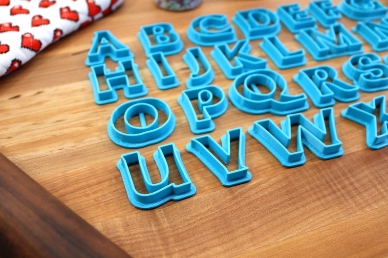 May include: A set of blue plastic alphabet cookie cutters on a wooden surface. The cutters are arranged in alphabetical order.