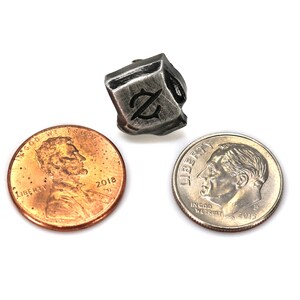 May include: A silver metal dice with a stylized "Z" symbol on each face. The dice is about the size of a US penny.