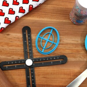 May include: A black ruler with a white knob in the center, a blue oval-shaped cookie cutter, a wooden rolling pin, a blue spatula, and a jar of sprinkles on a wooden surface.