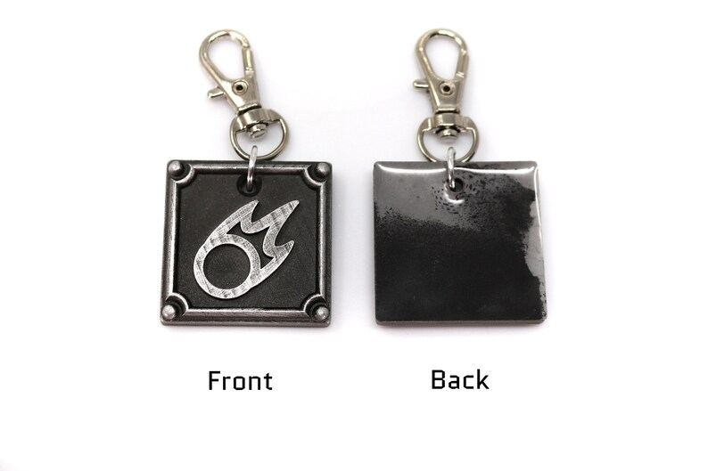 May include: A silver metal keychain with a black square charm featuring a silver flame symbol on the front and a blank surface on the back.