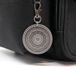 May include: A silver metal keychain with a circular charm featuring a geometric design and a triangle symbol in the center.