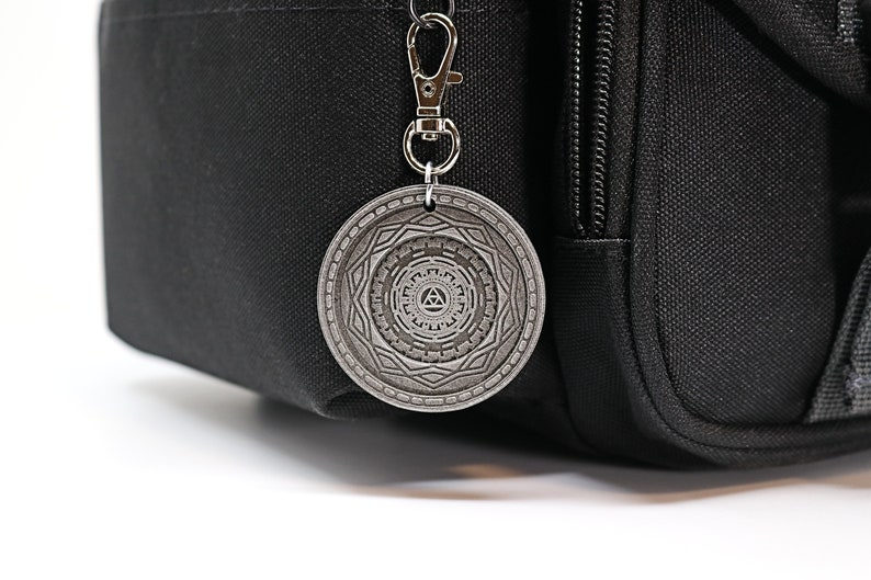 May include: A silver metal keychain with a circular charm featuring a geometric design and a triangle symbol in the center.