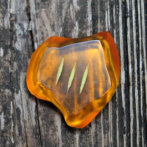 May include: A unique, amber-colored resin pendant with three vertical green lines. The pendant has a clear, slightly raised center and is set against a weathered wooden surface.