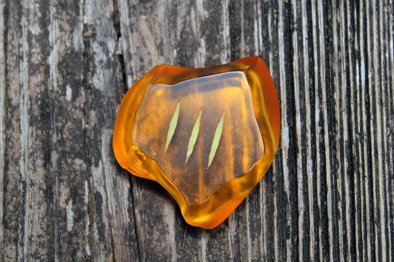 May include: A unique, amber-colored resin pendant with three vertical green lines. The pendant has a clear, slightly raised center and is set against a weathered wooden surface.