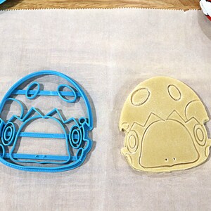 May include: A blue plastic cookie cutter and a light brown cookie in the shape of a cartoon character with a mushroom cap.