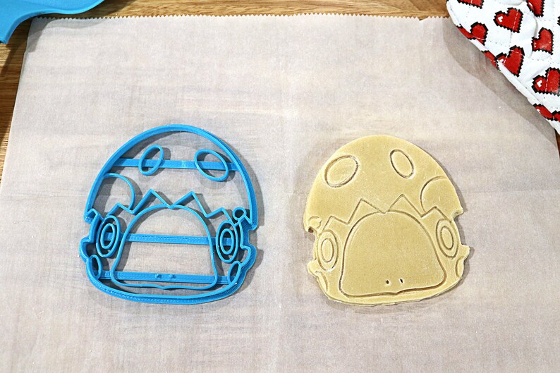 May include: A blue plastic cookie cutter and a light brown cookie in the shape of a cartoon character with a mushroom cap.