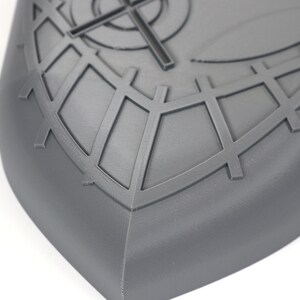 May include: A gray, shield-shaped object with a geometric design. The surface features a crosshair and grid pattern, with a rounded edge. The object is made of a solid material and is set against a white background.