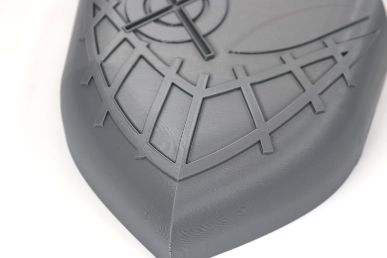 May include: A gray, shield-shaped object with a geometric design. The surface features a crosshair and grid pattern, with a rounded edge. The object is made of a solid material and is set against a white background.