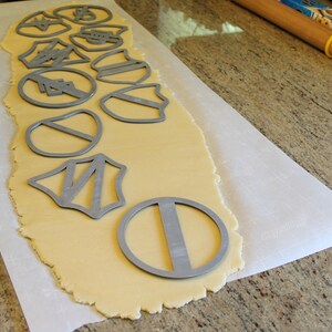 May include: A sheet of parchment paper with rolled out cookie dough and various metal cookie cutters on top. The cutters are in the shapes of stars, shields, and circles.