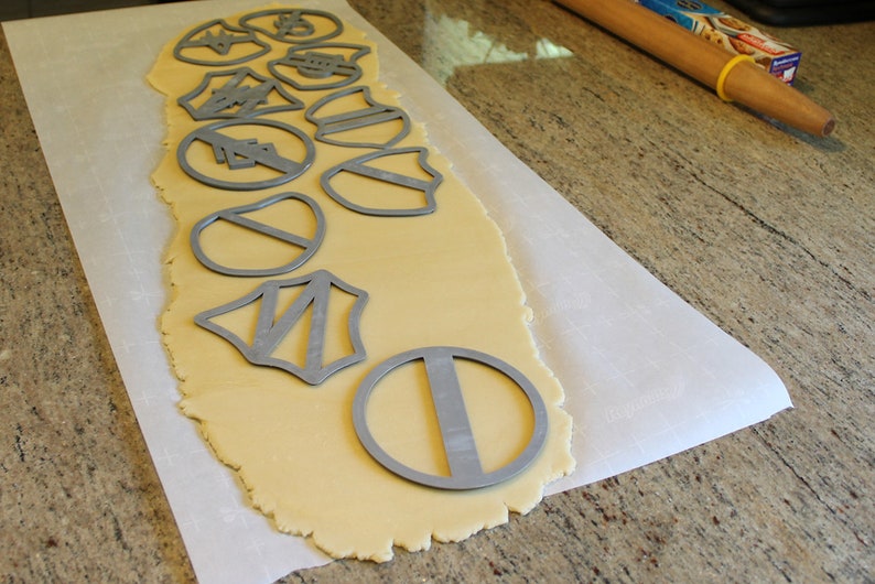 May include: A sheet of parchment paper with rolled out cookie dough and various metal cookie cutters on top. The cutters are in the shapes of stars, shields, and circles.