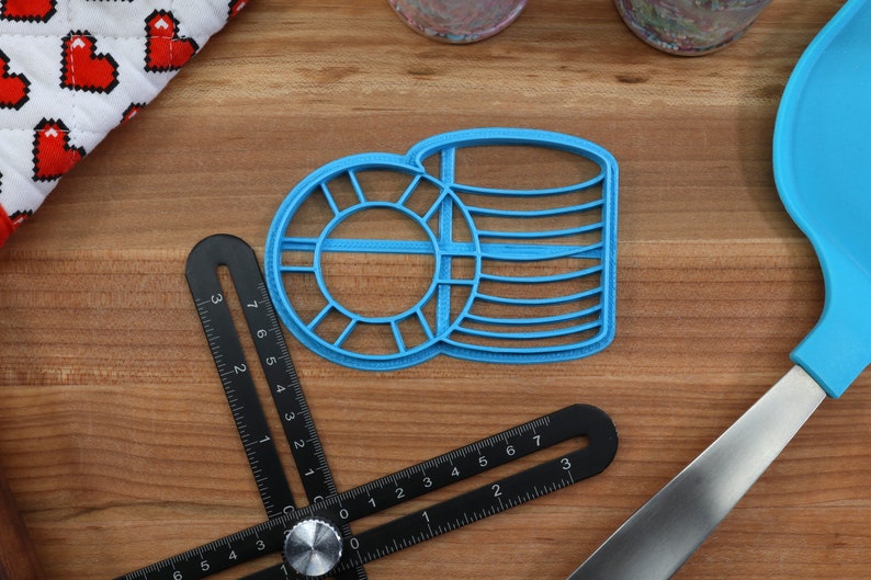 May include: Two blue plastic cookie cutters, one shaped like a circle with a smaller circle inside and the other shaped like a stack of coins. The cutters are on a wooden surface.