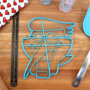 May include: Blue plastic cookie cutter shaped like a cartoon character with a hat and glasses. The cutter is on a wooden surface with a rolling pin, a ruler, and a blue spatula.