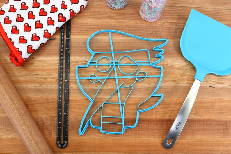 May include: Blue plastic cookie cutter shaped like a cartoon character with a hat and glasses. The cutter is on a wooden surface with a rolling pin, a ruler, and a blue spatula.
