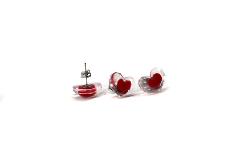 Puede incluir: Un par de pendientes transparentes en forma de coraz&oacute;n con corazones rojos en su interior. Los pendientes tienen postes de plata.