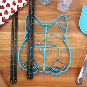 May include: A blue plastic cookie cutter shaped like a dog, with a heart-shaped nose, on a wooden surface with a rolling pin, a red oven mitt, and a blue spatula.