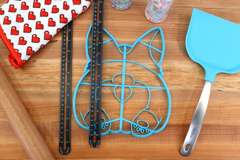 May include: A blue plastic cookie cutter shaped like a dog, with a heart-shaped nose, on a wooden surface with a rolling pin, a red oven mitt, and a blue spatula.