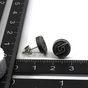May include: A pair of silver stud earrings with a black circular design featuring a stylised lightning bolt. The earrings are shown next to a ruler.