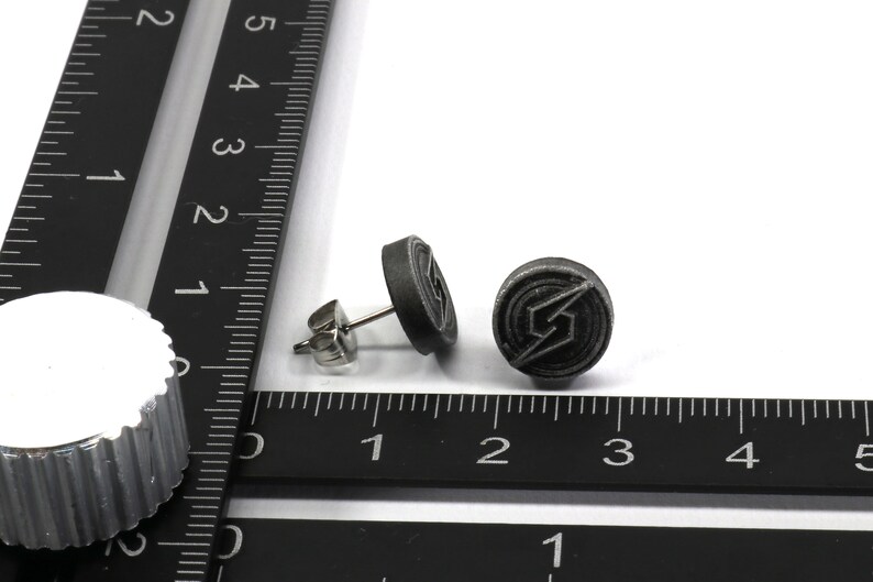 May include: A pair of silver stud earrings with a black circular design featuring a stylised lightning bolt. The earrings are shown next to a ruler.