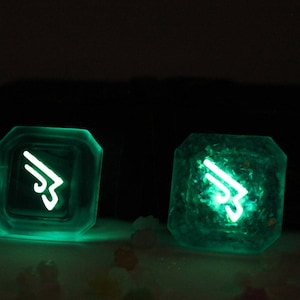 May include: Two glowing green resin dice with a white symbol on each face. The symbol is a stylized wing with a number 3 in the center.