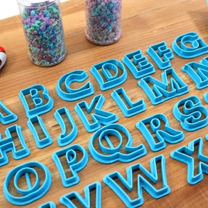 May include: A set of blue plastic alphabet cookie cutters on a wooden surface. The cutters are arranged in alphabetical order.