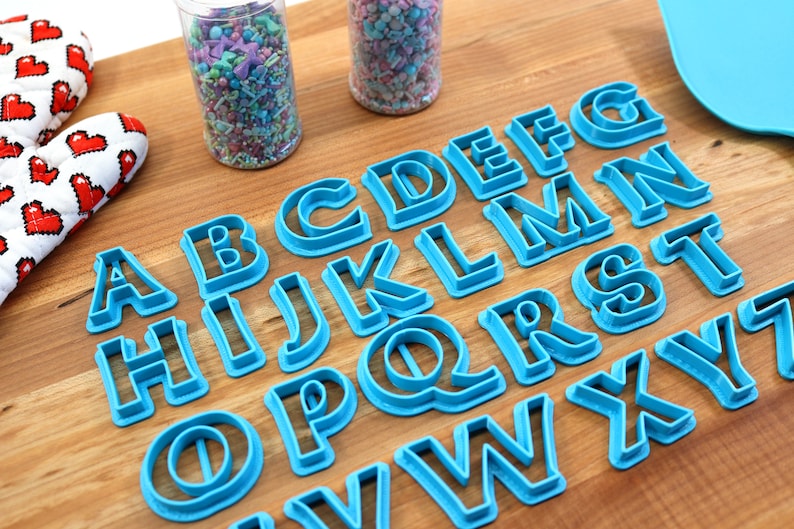 May include: A set of blue plastic alphabet cookie cutters on a wooden surface. The cutters are arranged in alphabetical order.