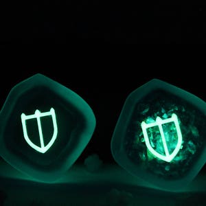 Puede incluir: Dos dados de resina verde oscuro con un s&iacute;mbolo de escudo blanco brillante en cada cara. Los dados se iluminan en la oscuridad.