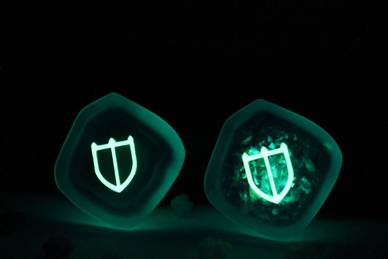 Puede incluir: Dos dados de resina verde oscuro con un s&iacute;mbolo de escudo blanco brillante en cada cara. Los dados se iluminan en la oscuridad.