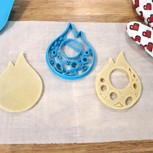 May include: Two cookie cutters in the shape of a flame, one blue and one made of dough. The blue cutter has a detailed design with circles and triangles. The dough cutter has a hole in the center.