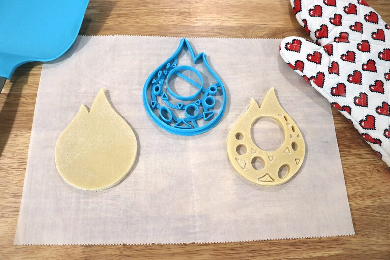 May include: Two cookie cutters in the shape of a flame, one blue and one made of dough. The blue cutter has a detailed design with circles and triangles. The dough cutter has a hole in the center.