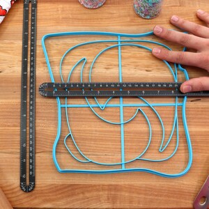 May include: A blue plastic baking mold shaped like a swirl design on a wooden cutting board. A ruler is placed on top of the mold to show the size.