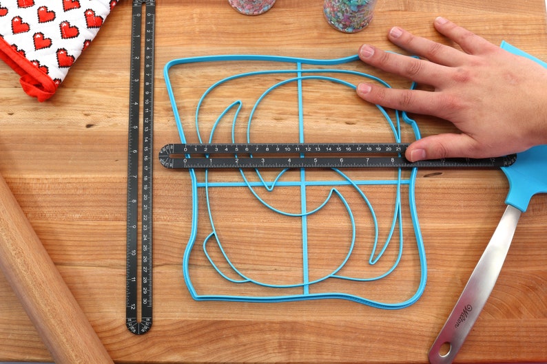 May include: A blue plastic baking mold shaped like a swirl design on a wooden cutting board. A ruler is placed on top of the mold to show the size.