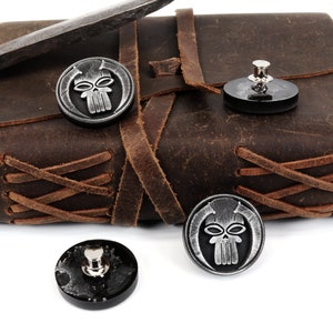 May include: Three silver skull-shaped buttons with black backgrounds. The buttons are on a brown leather journal with brown leather straps.