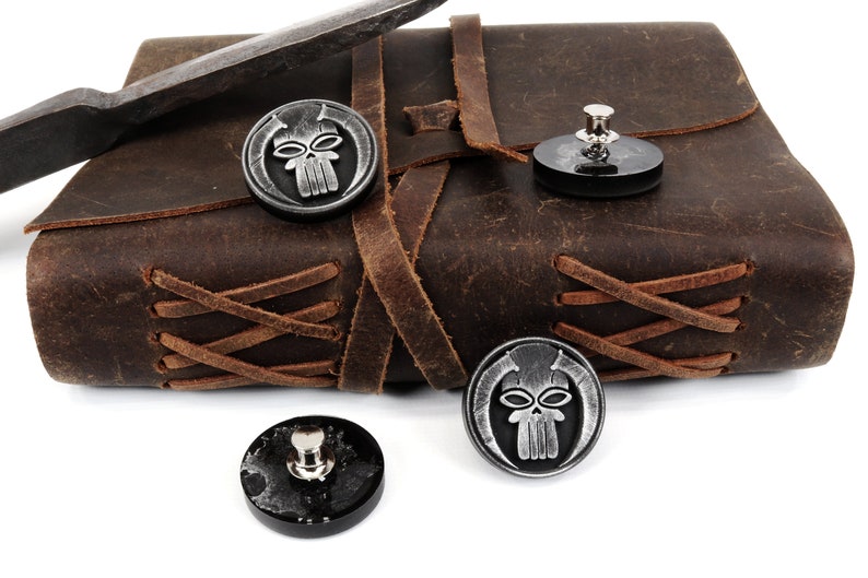 May include: Three silver skull-shaped buttons with black backgrounds. The buttons are on a brown leather journal with brown leather straps.