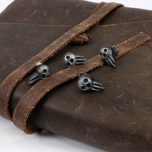 Peut inclure: Quatre fermoirs en m&eacute;tal en forme de cr&acirc;ne argent&eacute;s sur une sangle en cuir marron. Les fermoirs ont un design tridimensionnel et sont fix&eacute;s &agrave; la sangle par de petits rivets en m&eacute;tal.