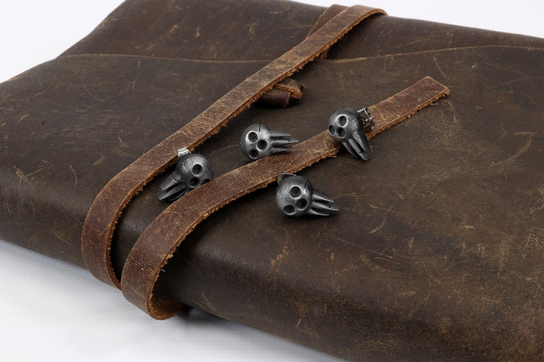 Peut inclure: Quatre fermoirs en m&eacute;tal en forme de cr&acirc;ne argent&eacute;s sur une sangle en cuir marron. Les fermoirs ont un design tridimensionnel et sont fix&eacute;s &agrave; la sangle par de petits rivets en m&eacute;tal.