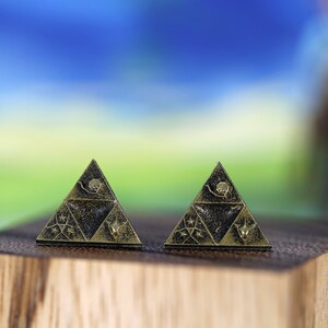 May include: Two gold-colored triangular earrings with a black background and intricate designs. The earrings are on a wooden surface.